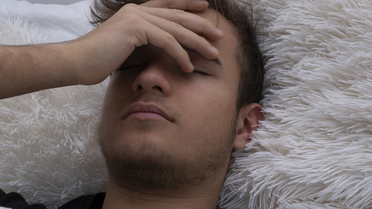 Junger Mann liegt erschöpft im Bett oder auf dem Sofa und hält sich eine Hand vor die Augen.
