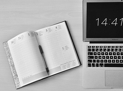 aufgeschlagener Terminkalender, daneben ein Laptop und eine digitale Zeitanzeige